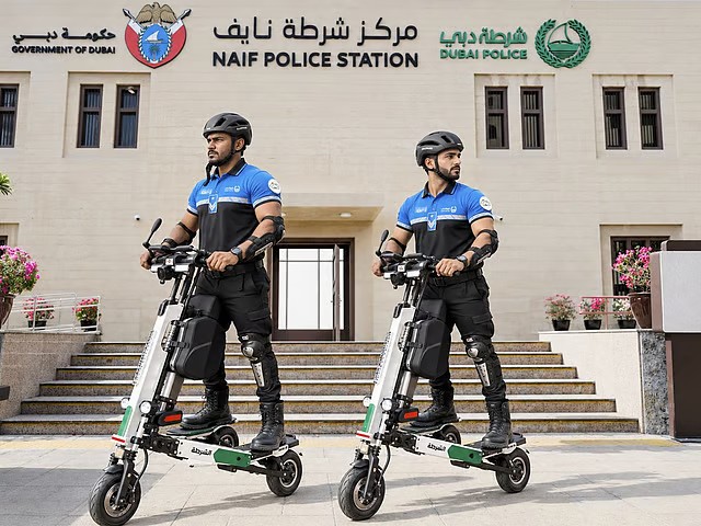 Two Dubai police officers on electric patrol scooters in front of Naif Police Station entrance wearing blue uniforms and helmets
