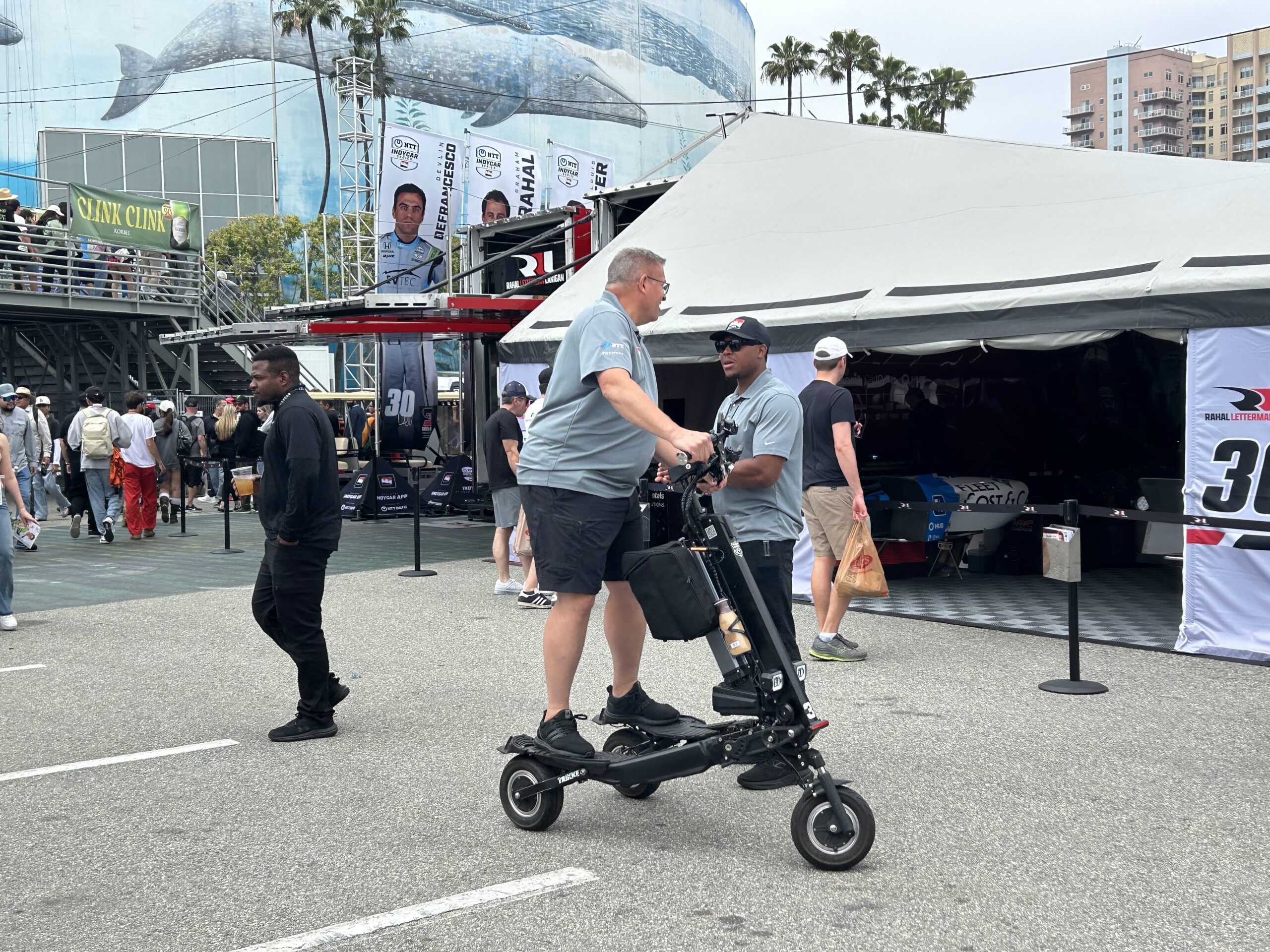 Two men in gray polo shirts ride small black racing scooters in a pit area at a motorsport event, with Acura/Indy branding in the background.