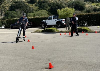 Man wearing a helmet rides a tall electric scooter through orange traffic cones in a parking lot with a security guard watching in the background