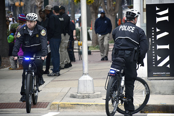 Two uniformed police officers ride bicycles with flashing blue lights on a city street, while pedestrians and a tall black banner appear in the background.