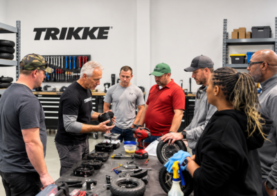 Group of people in a workshop examining motorcycle parts spread across a workbench in a TRIKKE garage setting