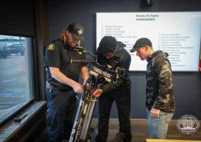 Three people including two Fargo Police officers inspect a mounted motorized device labeled Fargo Police in a briefing room with a whiteboard listing tasks