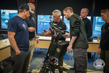 Group of men gathered around a bomb disposal robot on a wheeled platform as an instructor explains the controls