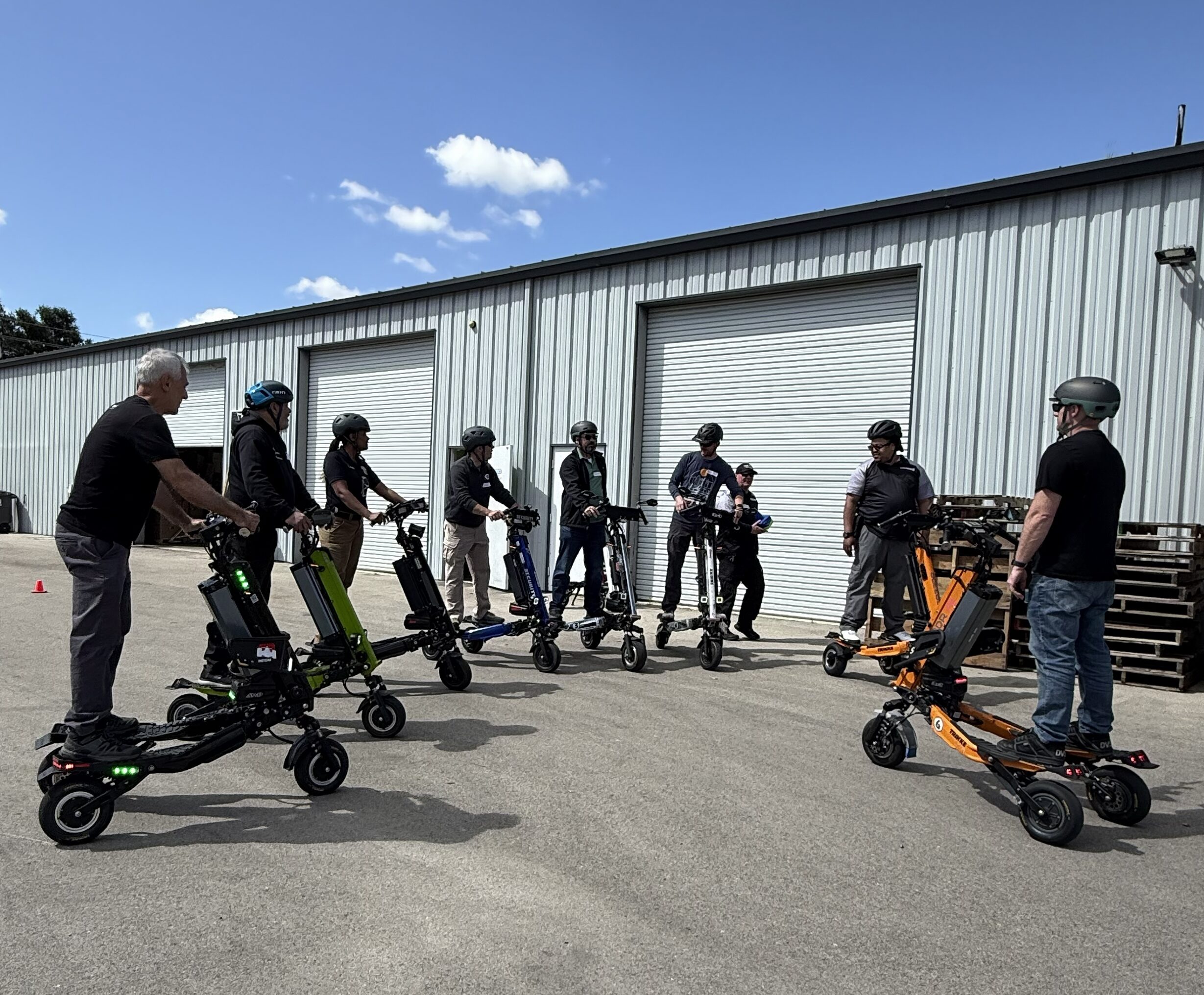 Group of people wearing helmets standing with electric scooters outside a metal warehouse